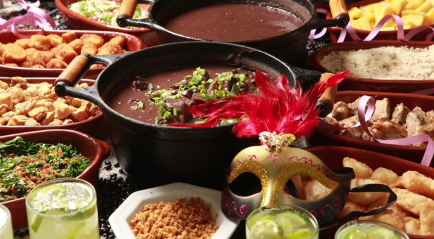 Foto de uma mesa com comidas de churrasco, duas panelas com feijoada no centro, copos de caipirinha, serpentinas e máscara