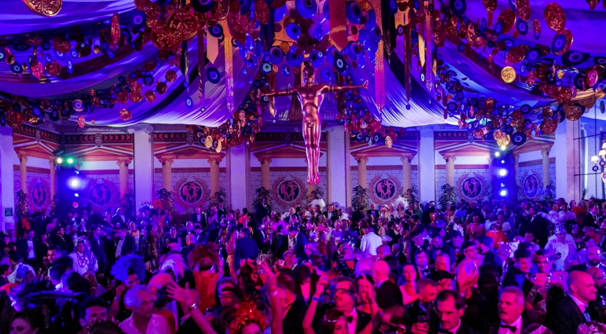 Foto de uma festa tipo baile, pessoas na pista, roupa estilo esporte fino, decorações no teto e uma decoração central grande.