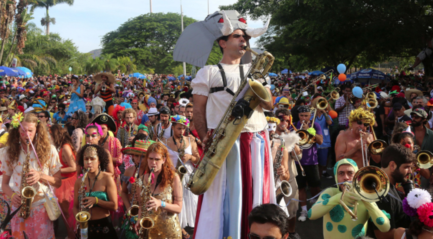 Foto de um dos integrantes do bloco de pernas de pau, tocando saxofone e fantasiado de elefante no meio da multidão no Aterro